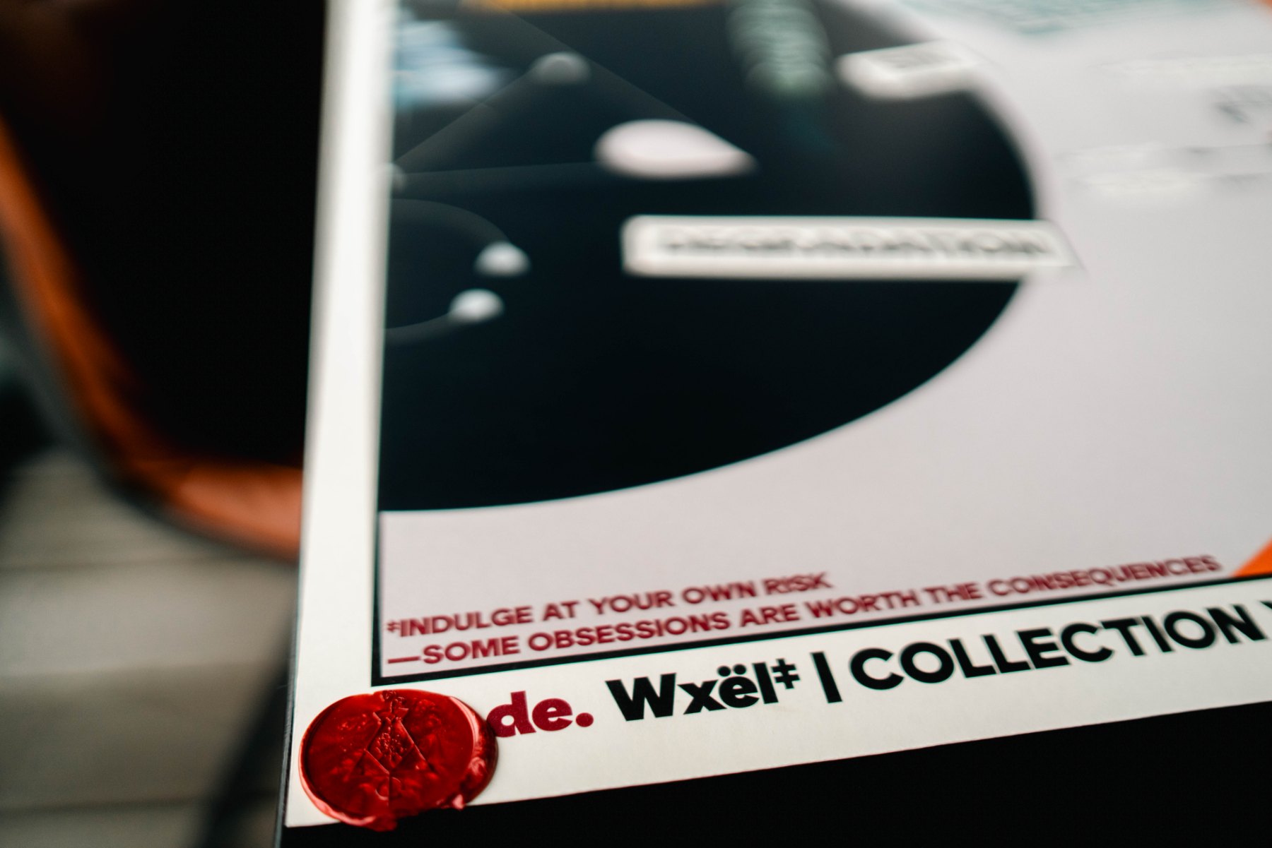 Bottom corner of the poster ; the INDULGE AT YOUR OWN RISK footnote, the de. Wxël colophon, and the red wax seal