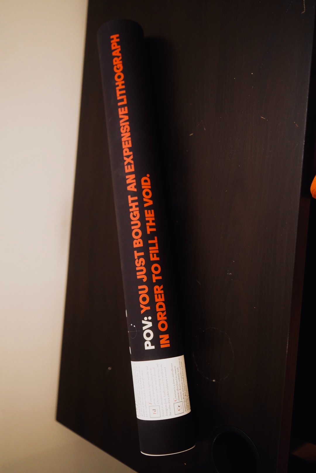 The black postal tube on dark wood, printed lengthwise with POV: YOU JUST BOUGHT AN EXPENSIVE LITHOGRAPH IN ORDER TO FILL THE VOID